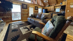 Interior acogedor de una cabaña con techo de madera, un sofá, una silla, un tapete con estampados de animales y una zona de cocina.