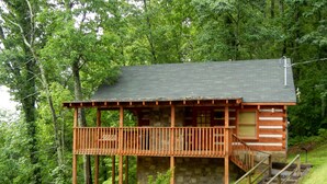Una cabaña con terraza de madera y base de piedra, rodeada de un bosque denso.