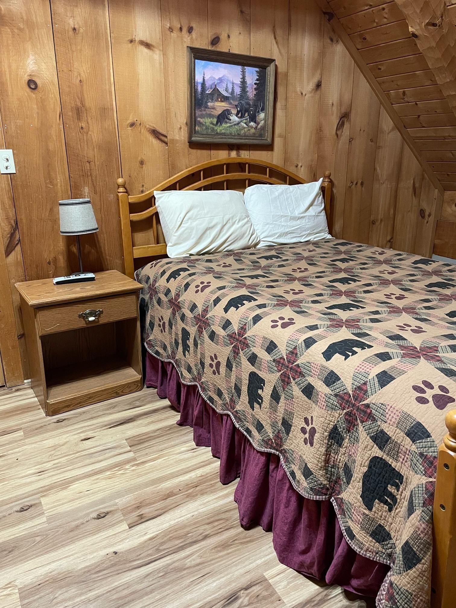 A wooden-paneled room with a bed, a nightstand, a lamp, and a painting on the wall.