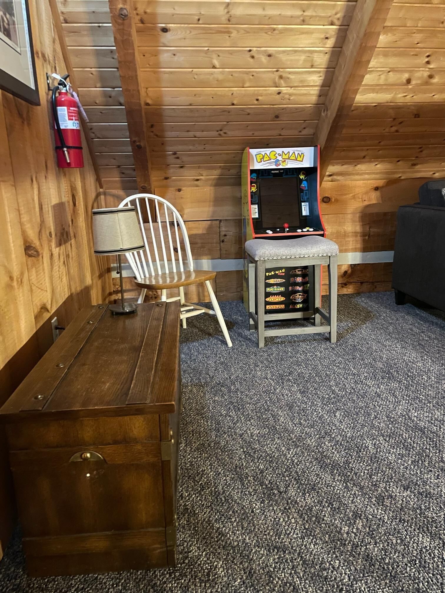 A wooden room with a wooden ceiling, a fire extinguisher, a Pac-Man arcade machine, a white chair, and a small table.