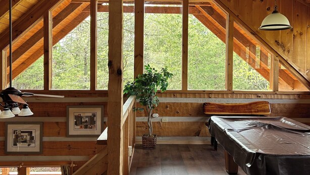 Une vaste pièce mansardée avec un plafond en pente en bois, de grandes fenêtres, une table de billard et un ventilateur de plafond.