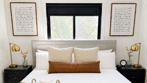 A neatly made bed with a brown accent pillow, a woven basket, and a green throw blanket. There are two bedside tables with lamps, and the room has a large window with a view of trees. - Modern home with privacy of the hills right in town! (Rapid City)