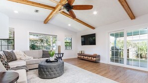 Ein geräumiges Wohnzimmer mit Deckenventilator, großen Fenstern, einer Couch, einem Hocker und einem an der Wand befestigten Fernseher.