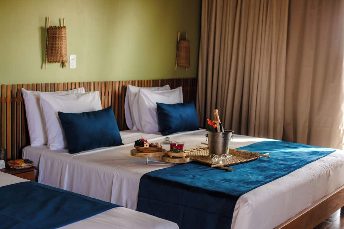 Quarto de hotel com duas camas, cada uma com roupa de cama branca e detalhes azuis. Há uma bandeja de café da manhã na cama com frutas, uma bebida e uma cesta.