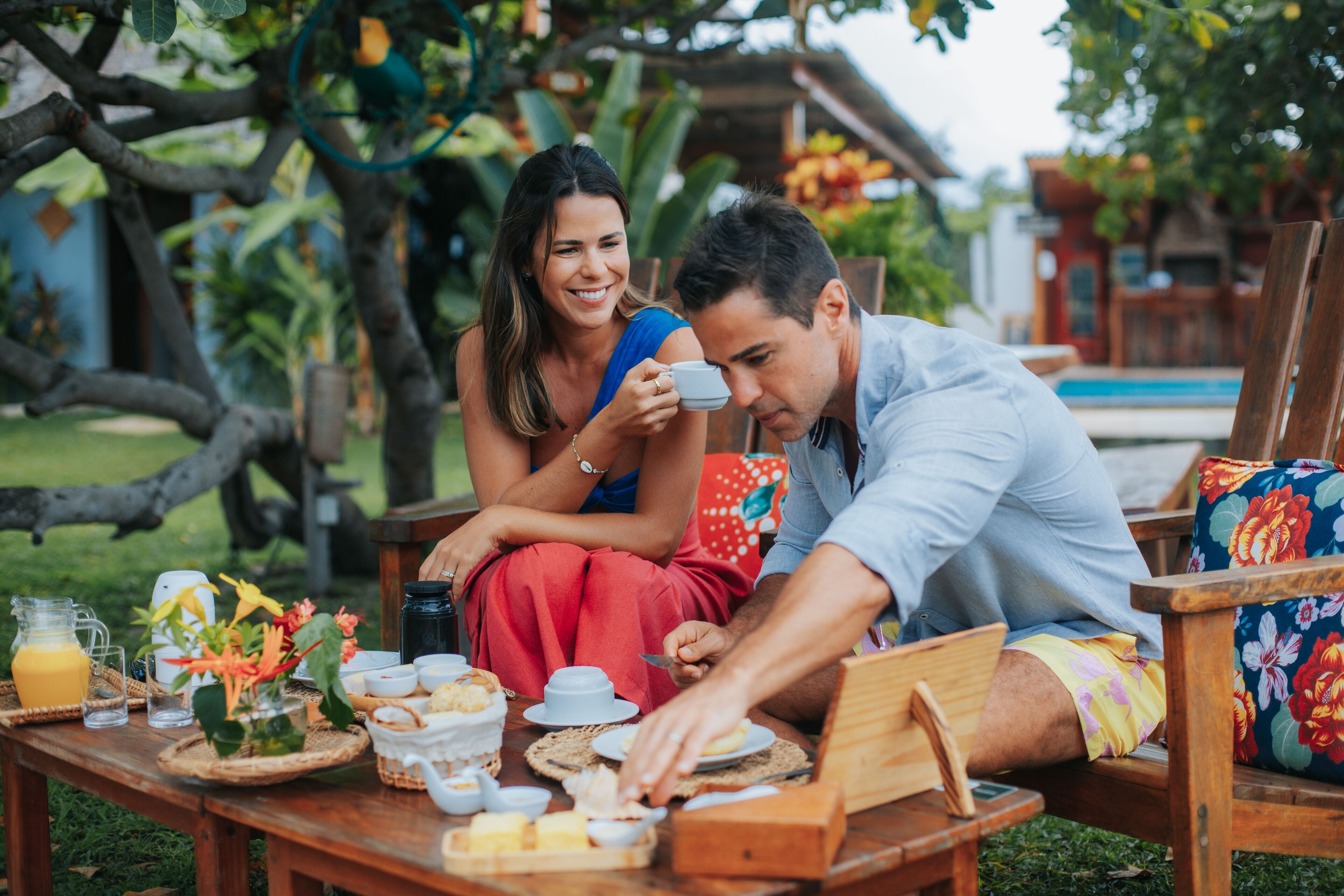 Una pareja cenando al aire libre, con una mesa dispuesta para el desayuno, rodeada de exuberante vegetación y una piscina al fondo.