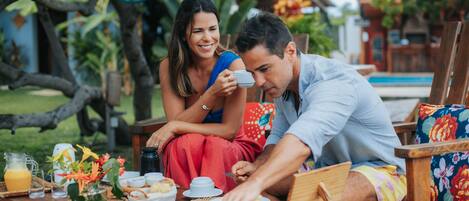 Una pareja cenando al aire libre, con una mesa dispuesta para el desayuno, rodeada de exuberante vegetación y una piscina al fondo.
