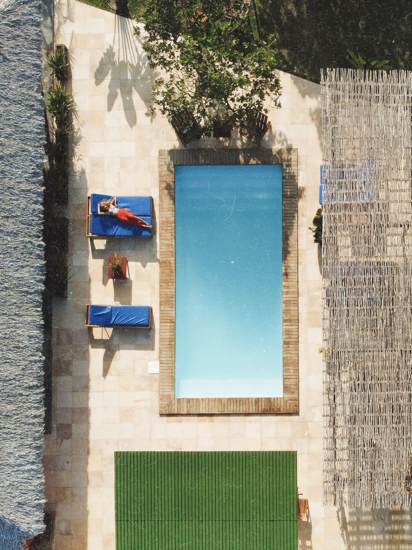 An aerial view of a swimming pool with two sun loungers and a green mat, surrounded by a wooden deck and a bamboo fence.