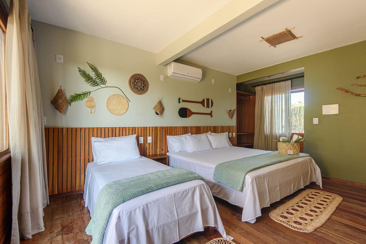 Quarto com duas camas, painéis de madeira, tapete tecido e decorações nas paredes, incluindo uma samambaia e um objeto circular tecido.