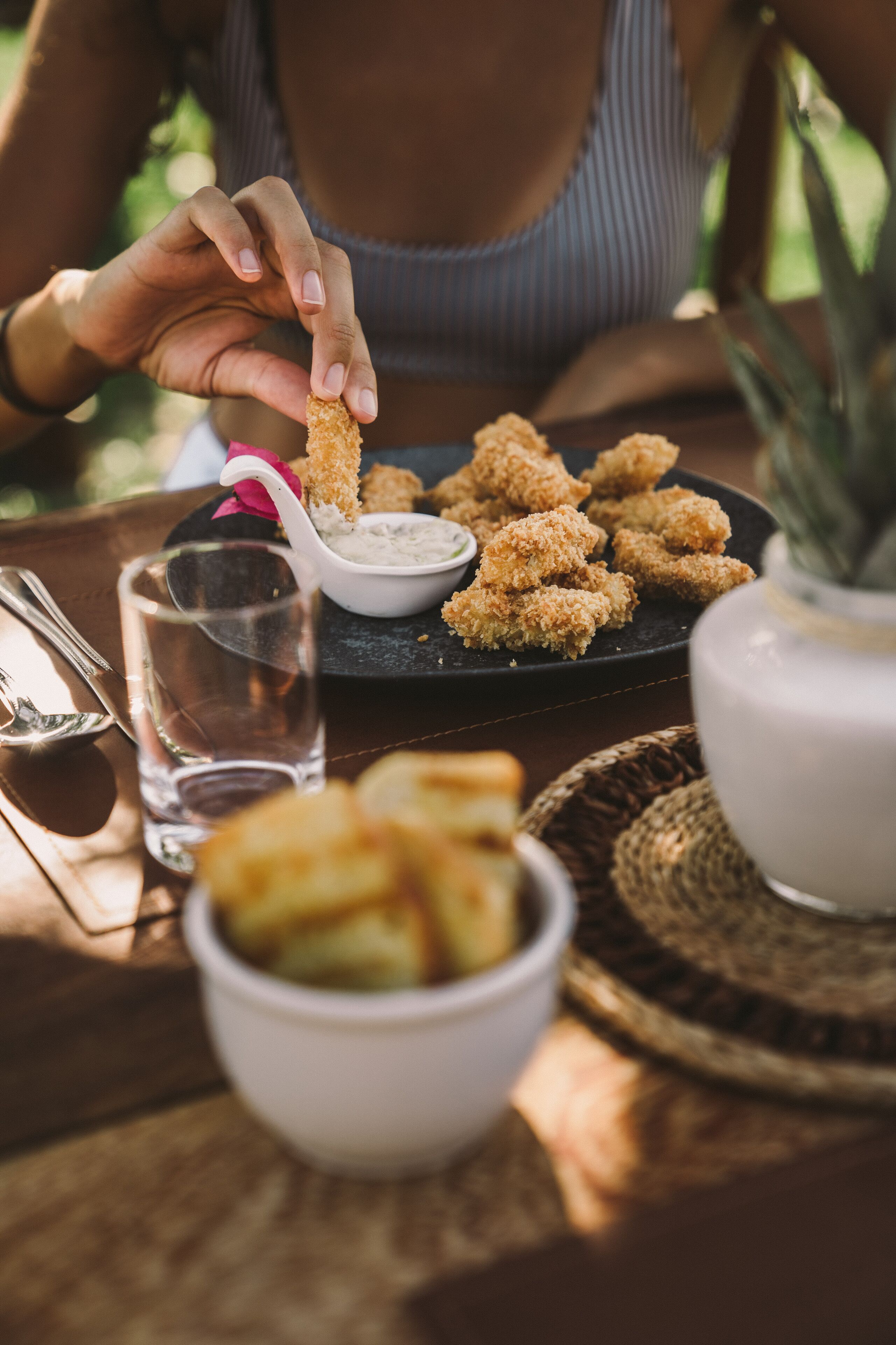 A person holding a fried item, with a plate of similar items and a bowl of dipping sauce on a table.