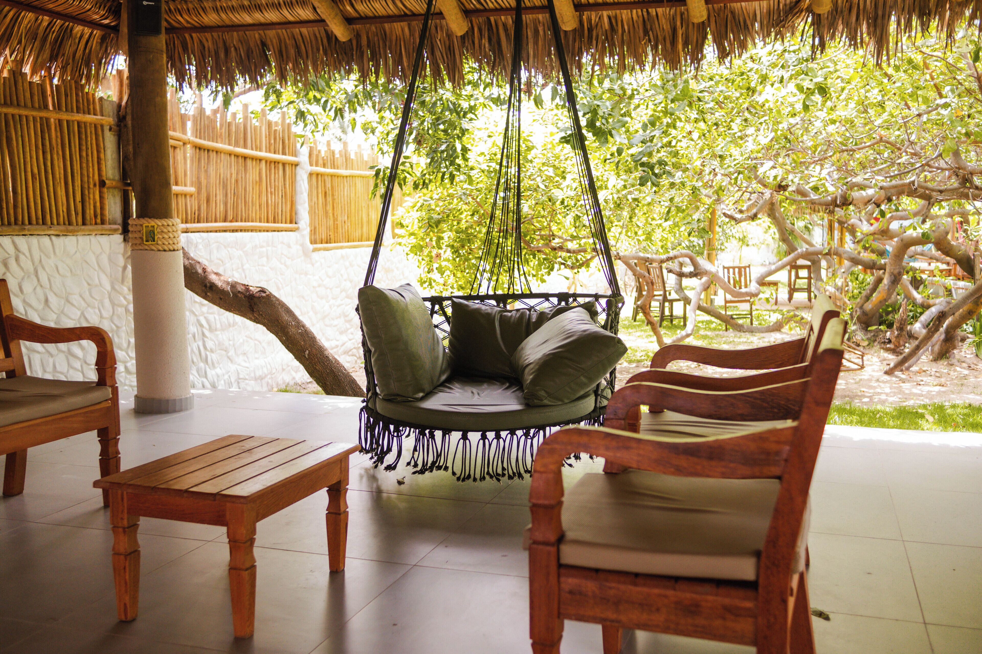 A wooden swing chair with cushions, a wooden table, and chairs in a shaded outdoor area with a thatched roof.