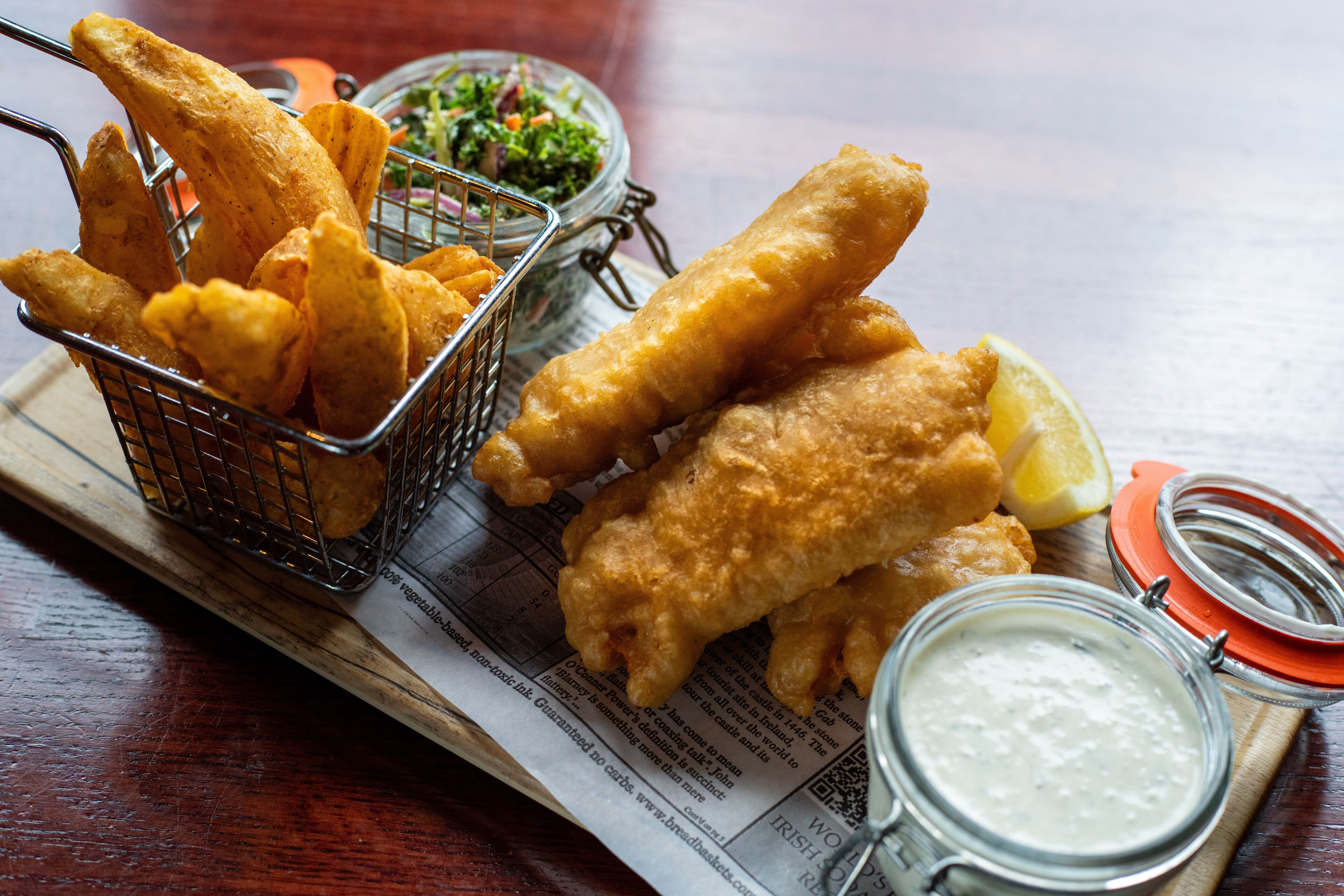 A basket of fried fish and chips, a lemon wedge, and a small jar of sauce on a newspaper.