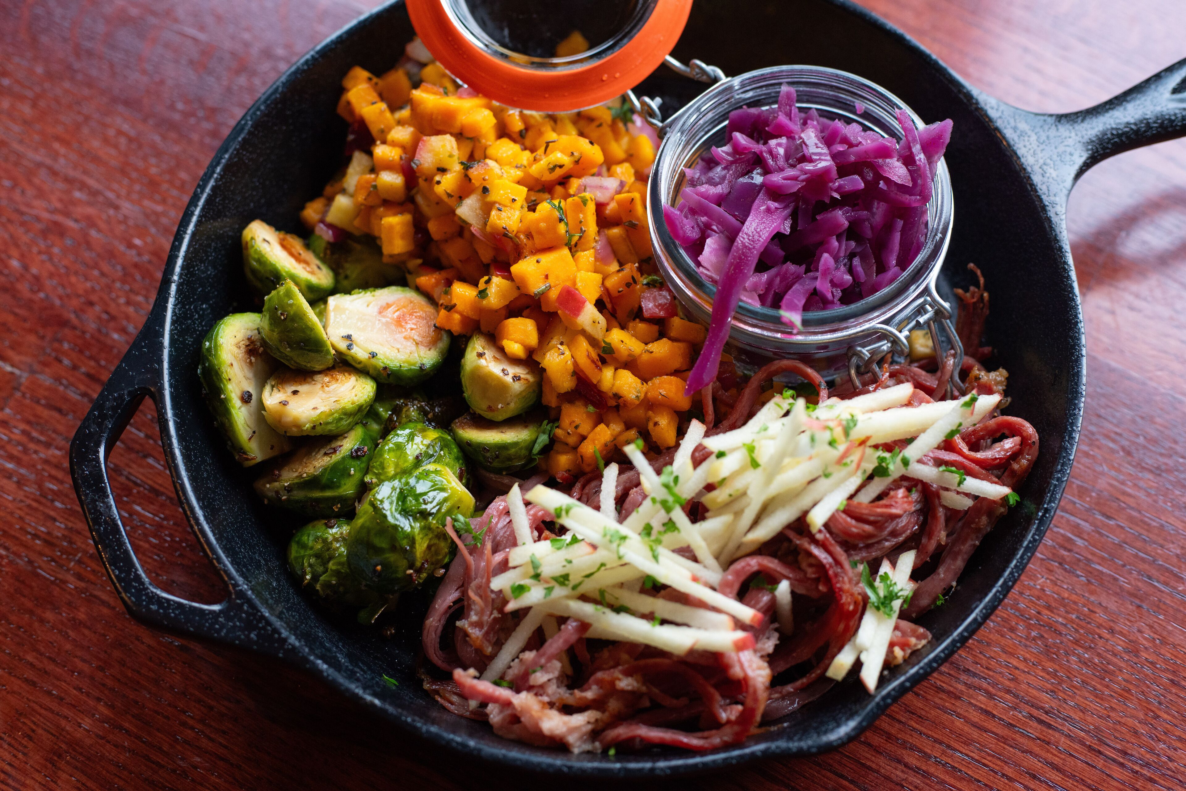 A cast iron skillet filled with various cooked vegetables and meats, including Brussels sprouts, carrots, and red onions, with a small jar of pickled red onions on the side.