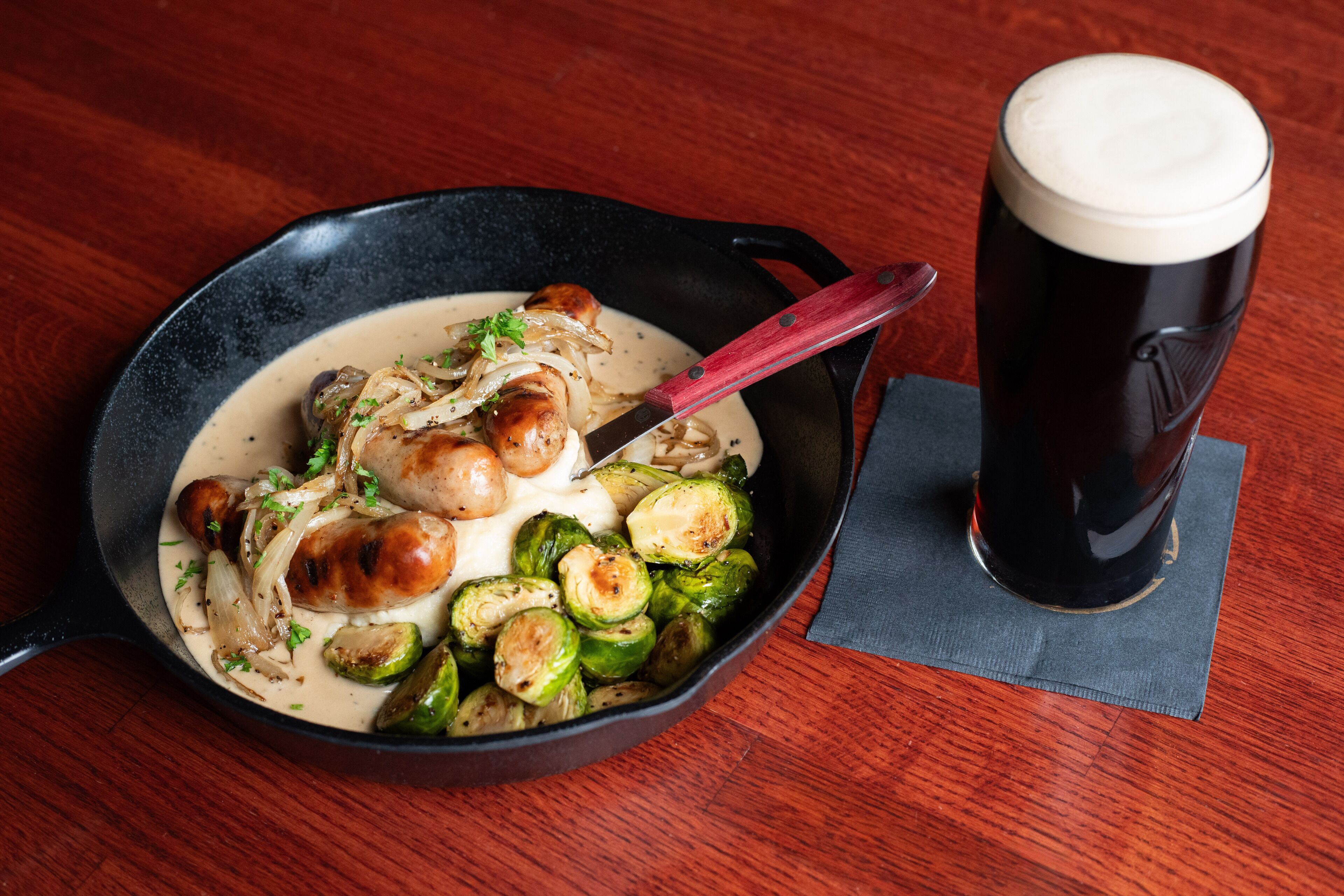 Un plat amb botifarra i brussel·les, i un got de cervesa damunt una taula de fusta.