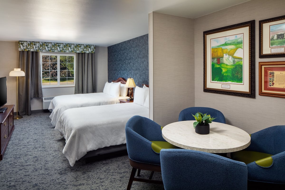 A hotel room with two beds, a round table with chairs, a TV, and framed artwork on the wall.