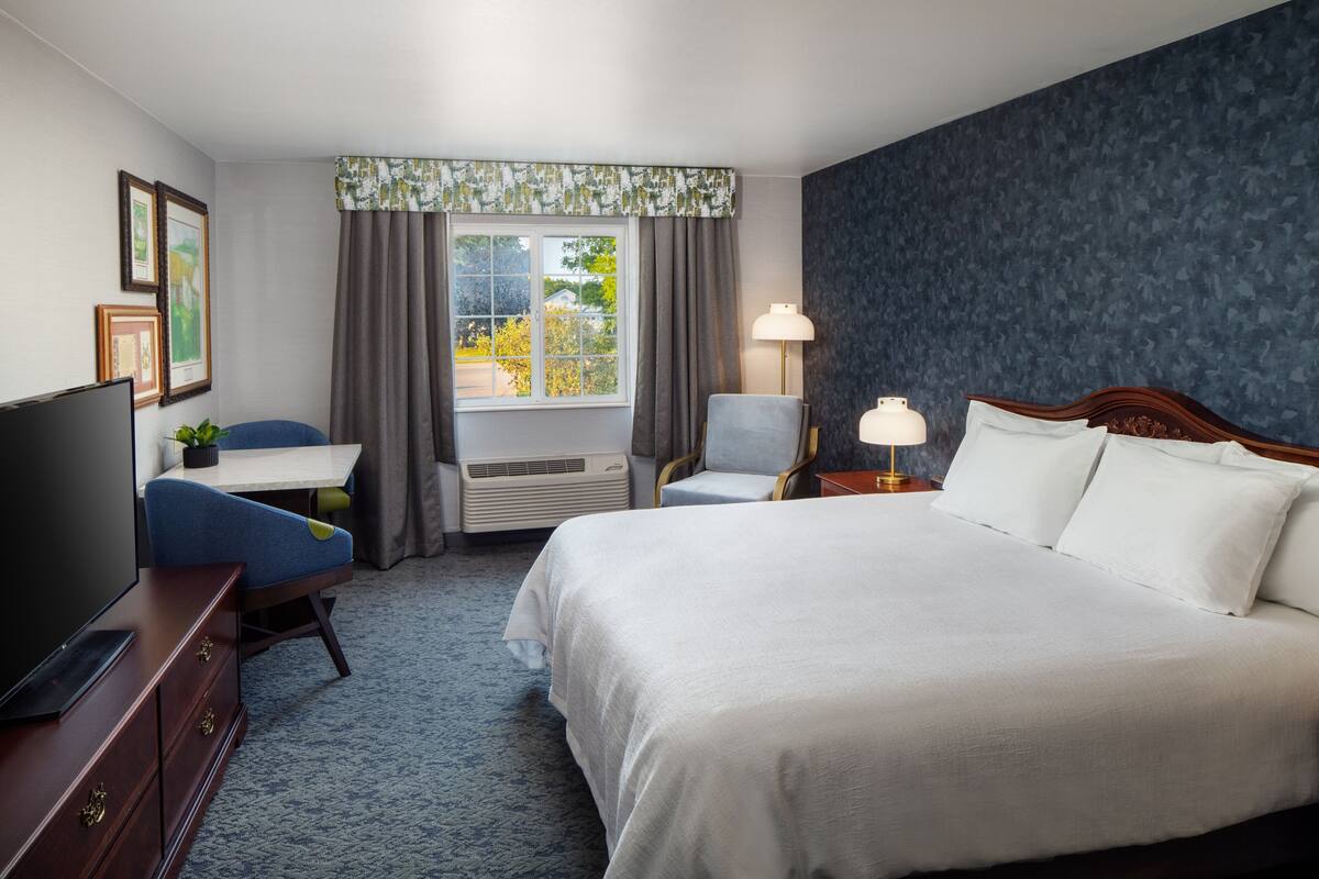 A hotel room with a large bed, a desk with a chair, a TV, and a window with curtains.