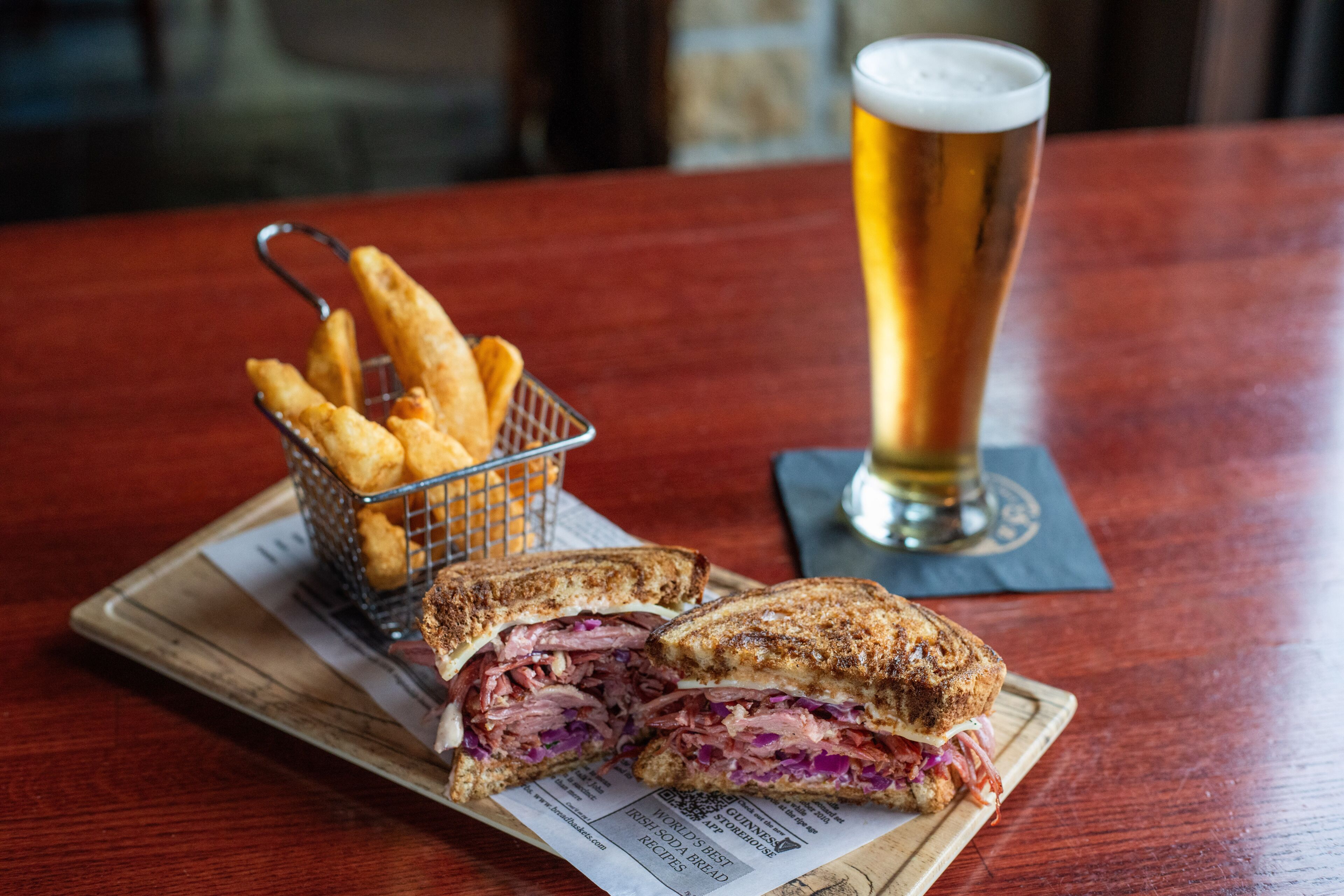 A sandwich cut in half, a glass of beer, and a basket of fries on a wooden table.