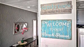 A reception area with a 'Welcome to our Beach House' sign, a 'Sandy toes & salty kisses' sign, and a decorative mirror with a floral arrangement. - Beach Service|Gulf Front|Private Balcony|PCB|CC514 (Panama City Beach)