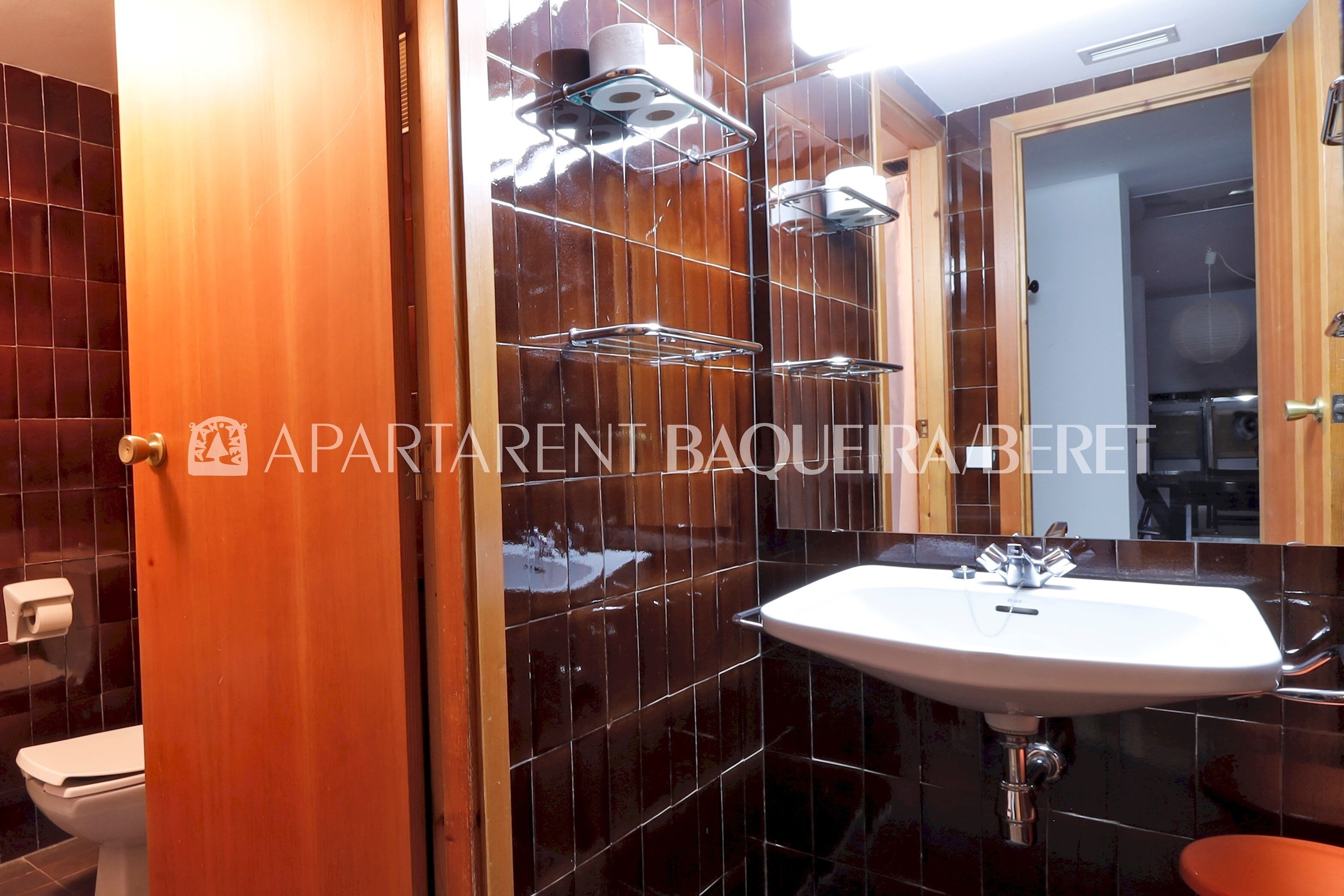 Un baño con un lavabo blanco, un espejo y un inodoro. Hay dos repisas sobre el lavabo, y las paredes están cubiertas de azulejos.
