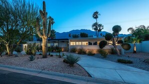 Una casa moderna con un jardín desértico, que incluye cactus y palmeras, y una vista de la montaña al fondo.