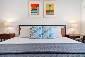 A hotel room with a large bed, two nightstands, two wall-mounted lamps, and two framed artworks depicting Palm Springs.