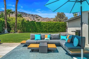 A modern outdoor living area with a gray sofa, wooden coffee table, and a blue umbrella.