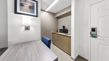 A hotel room with a dining table, a blue chair, a kitchen area with a coffee machine, and a framed picture on the wall.