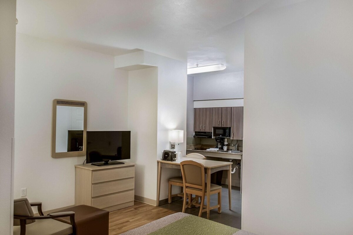 Una habitación pequeña con cama, escritorio, silla, televisor, espejo y kitchenette.
