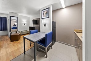 A compact hotel room with a kitchenette, a dining area with a table and chairs, a flat-screen TV mounted on the wall, and a refrigerator. - Quality Inn Fishers - Indianapolis (Fishers)