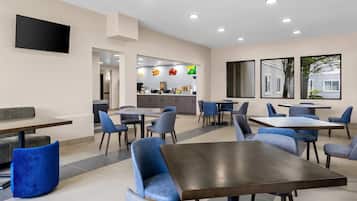 A modern dining area with multiple tables and chairs, a kitchen in the background, and a flat-screen TV mounted on the wall.