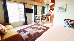 A room with a bed, a sofa, a dining table with chairs, a TV mounted on the wall, and a staircase leading to another level.