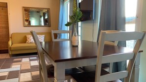 A dining area with a wooden table and white chairs, a green sofa with patterned cushions, a mirror, and a TV mounted on the wall.