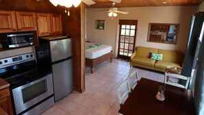 A compact kitchen with a microwave, stove, and refrigerator. There is a dining table with chairs and a ceiling fan. A sofa and a window with a view of the outdoors are also visible.