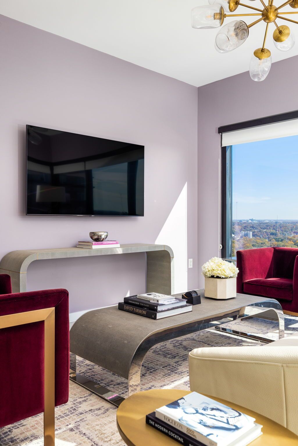 Ein modernes Wohnzimmer mit Flachbildfernseher, einer geschwungenen Konsole, roten Sesseln und Blick auf eine Stadtlandschaft durch große Fenster.