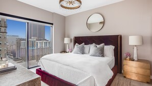 Ein modernes Hotelzimmer mit einem großen Bett, zwei Nachttischlampen, einem runden Spiegel und Blick auf die Stadt durch raumhohe Fenster.