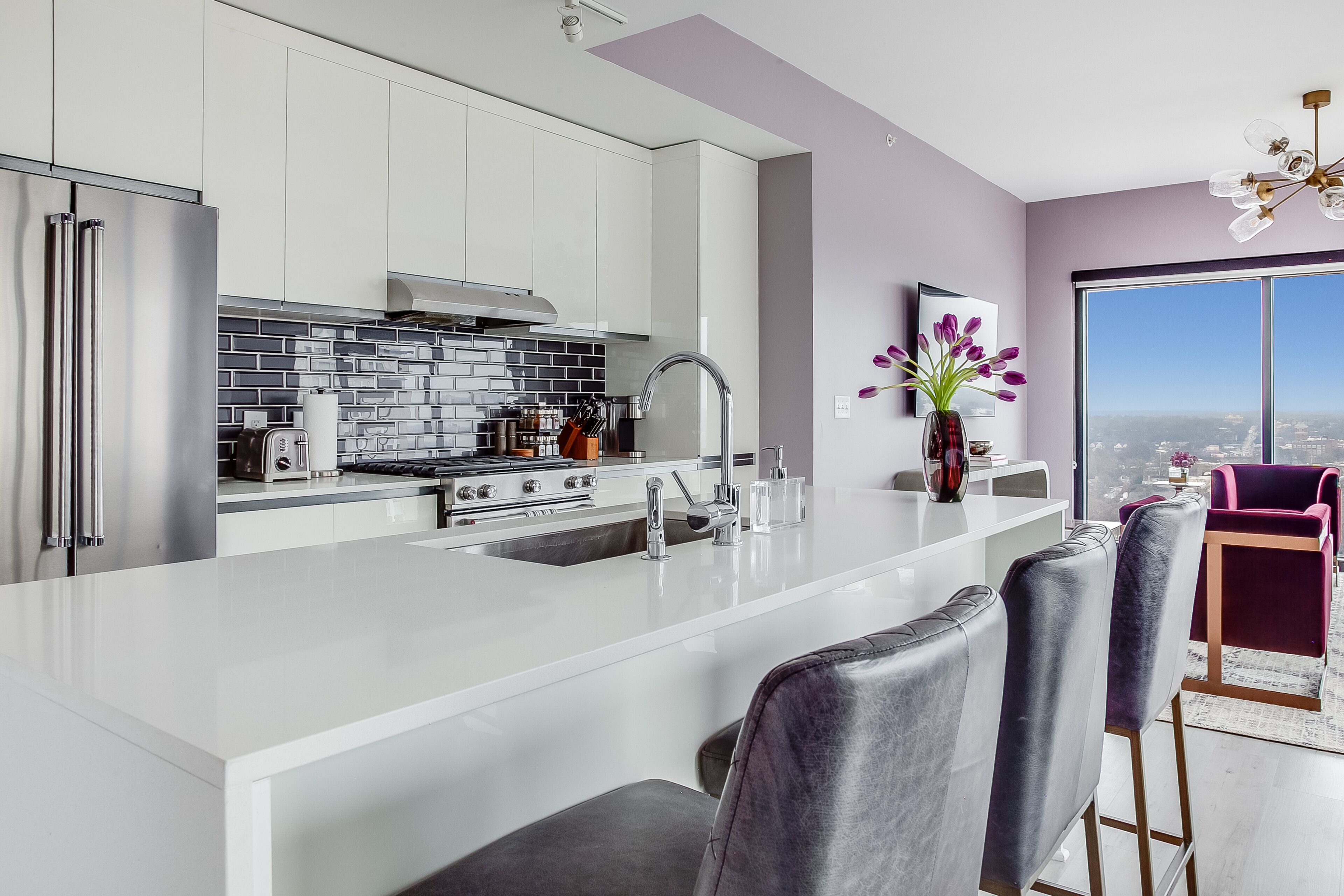 A modern kitchen with white cabinets, a large island, and a view of the city through a large window.