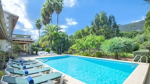 A rectangular swimming pool with clear blue water, surrounded by lounge chairs with blue towels, palm trees, and a mountainous backdrop.