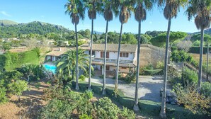 A house with a tiled roof, surrounded by palm trees, a swimming pool, and a hilly landscape.