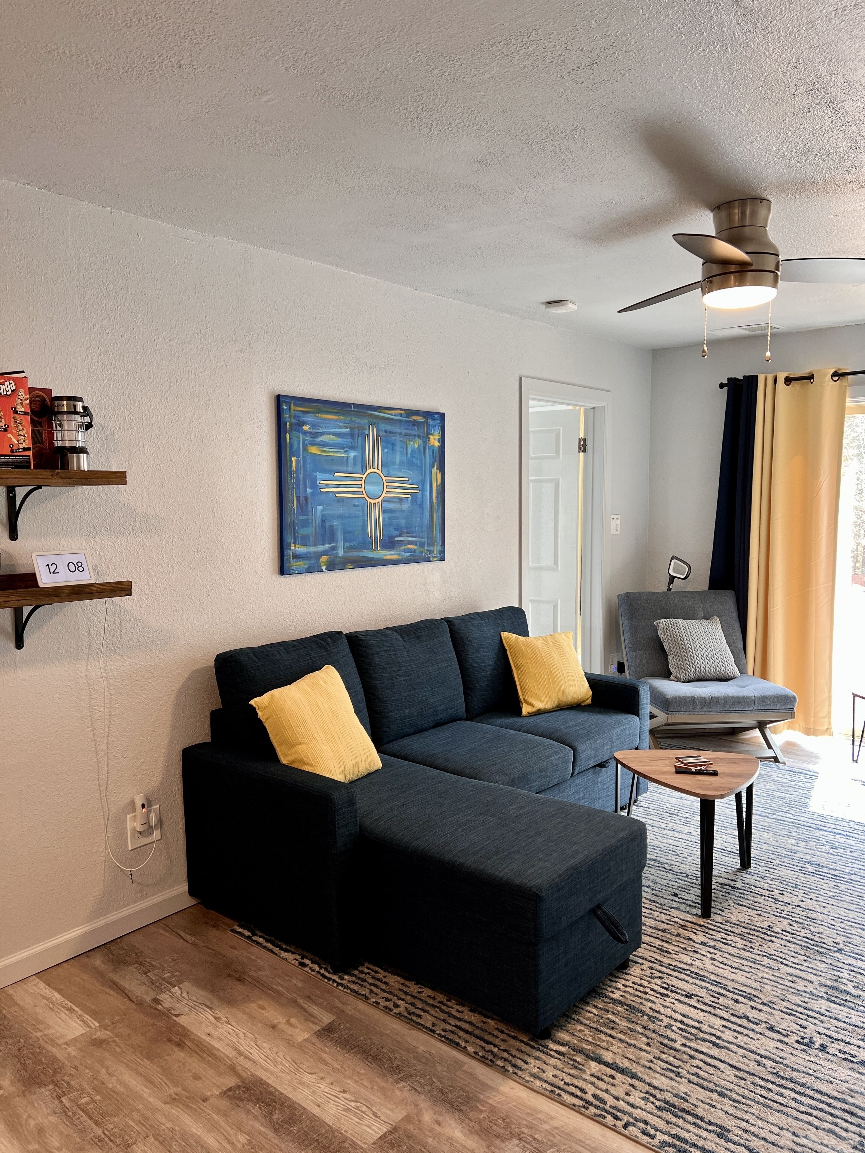 A modern living room with a dark grey sectional sofa, a small wooden coffee table, and a grey armchair. There is a ceiling fan, a blue abstract painting, and a door with a white frame.