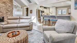 Ett modernt vardagsrum med en lädersoffa, en grå fåtölj och en flätad pall. I bakgrunden syns ett kök med vita skåp och en matplats med blå barstolar.