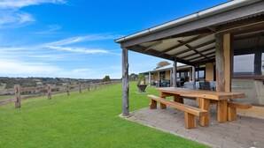 A covered outdoor seating area with a wooden table and benches, overlooking a grassy field and a vineyard.