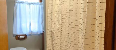 Et badeværelse med toilet, bruseforhæng og et vindue med gardin.