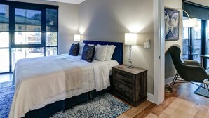 Ein Schlafzimmer mit einem großen Bett, zwei Nachttischlampen, einem Nachttisch, einem Sessel und Blick durch große Fenster nach draußen.