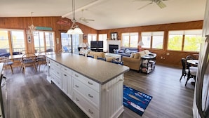 A spacious kitchen with a large island, wooden flooring, and a dining area with a table and chairs. There are ceiling fans, a fireplace, and large windows providing natural light.