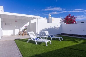 A white building with a covered outdoor seating area, artificial grass, and a red flowering plant. - Rooms & Suites Leticia, Puerto del Carmen (Tías)