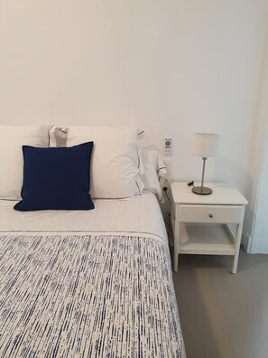 Un llit ben fet amb un edredó estampat, una almoina blava i almoines blanques. Hi ha una tauleta de nit blanca amb una làmpada i una presa corrent.