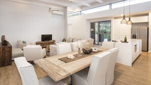 Ein modernes Wohnzimmer mit einem Holzespeisetisch, weißen Polsterstühlen, einem Fernseher und einer Küchenzeile mit Kühlschrank und Pendelleuchten.