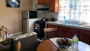A compact kitchen with a refrigerator, stove, and wooden cabinets. A dining table with chairs is in the foreground, and a TV is mounted on the wall.