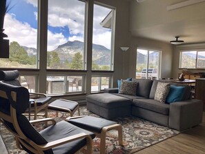 Un salon spacieux avec un canapé d’angle gris, deux fauteuils confortables et une grande fenêtre donnant sur une vue imprenable sur les montagnes.