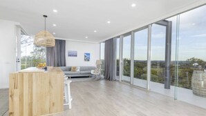 A spacious living area with a wooden coffee table, a gray sofa, and a rocking chair. Large windows offer a view of the outdoors.