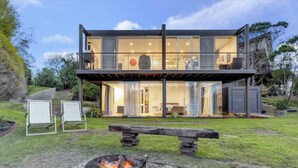 A modern two-story house with large glass windows, a balcony, and a fire pit in the yard.